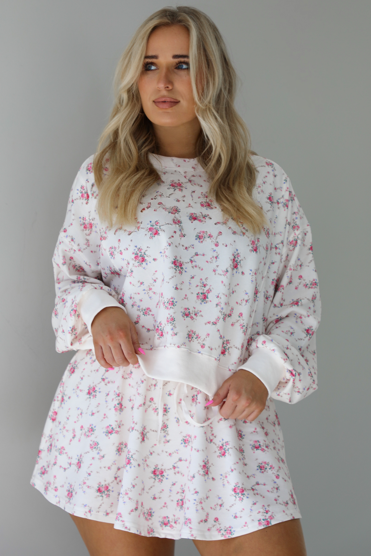 Woman wearing a pale pink two piece set that has a pink floral print pattern throughout. The set featuring an oversized long sleeved top and high waisted skirt. The photo is taken indoors against a white wall.