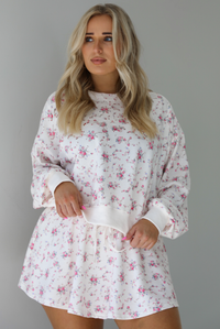 Woman wearing a pale pink two piece set that has a pink floral print pattern throughout. The set featuring an oversized long sleeved top and high waisted skirt. The photo is taken indoors against a white wall.