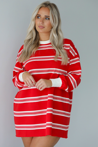 Woman wearing a red with white stripes long sleeved t-shirt dress. The photo is taken indoors against a white wall.