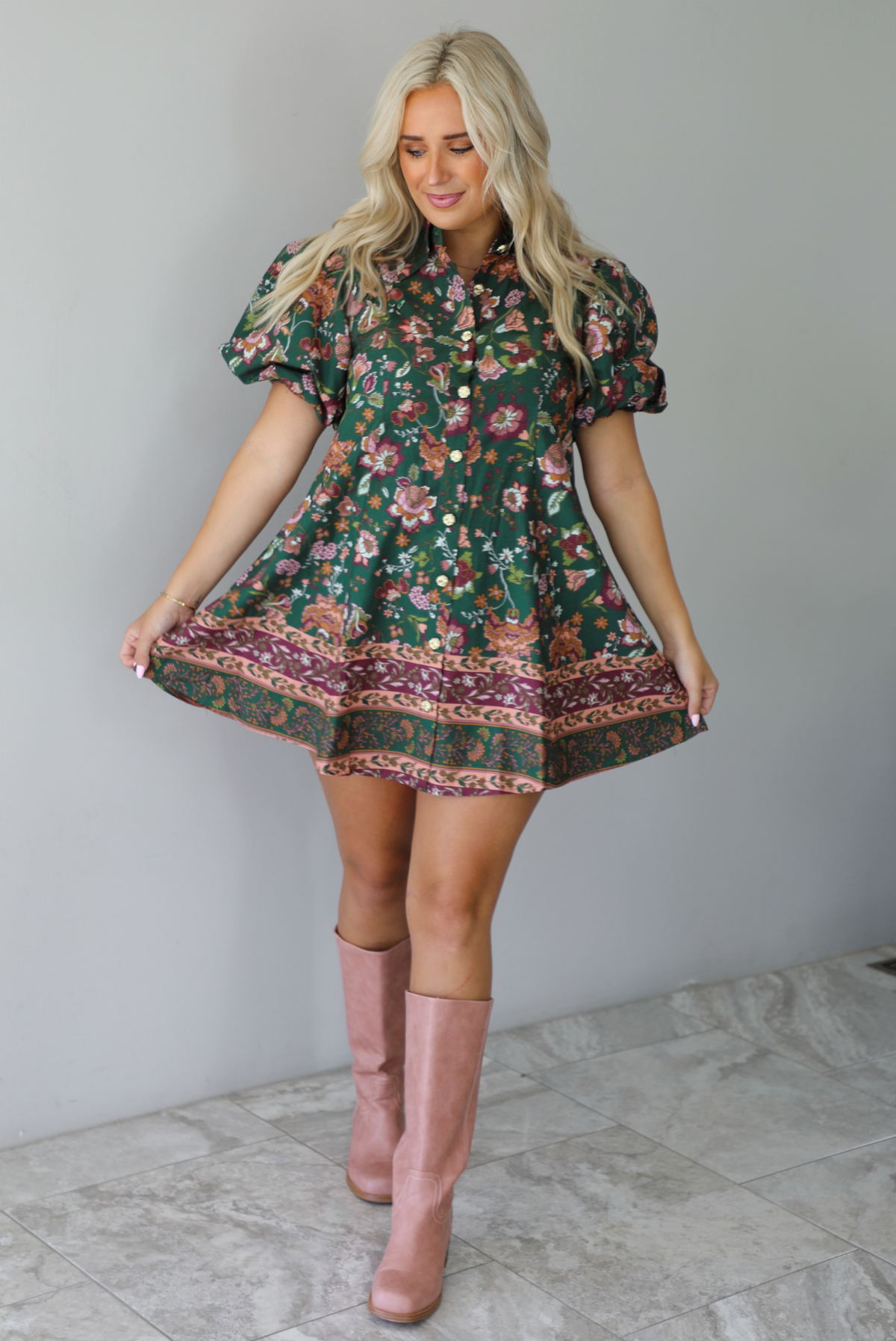 A woman wearing a dark green dress that has a mauve and multicolored floral print pattern throughout, has puffed sleeves, a collared neckline with functional gold button closures in the front, paired with muted pink boots, standing indoors against a white wall with white marble flooring. 