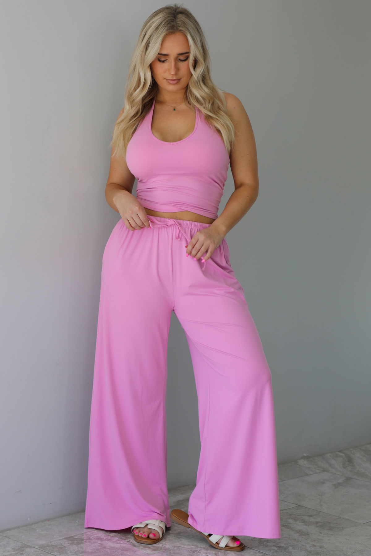 Woman wearing a two pieced pink set featuring a halter top and comfy pants with a flared hem detail. The photo is taken indoors against a white wall with white marble flooring. 