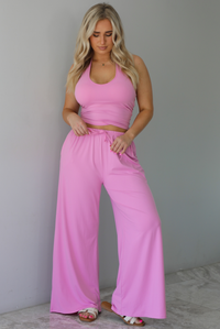 Woman wearing a two pieced pink set featuring a halter top and comfy pants with a flared hem detail. The photo is taken indoors against a white wall with white marble flooring. 