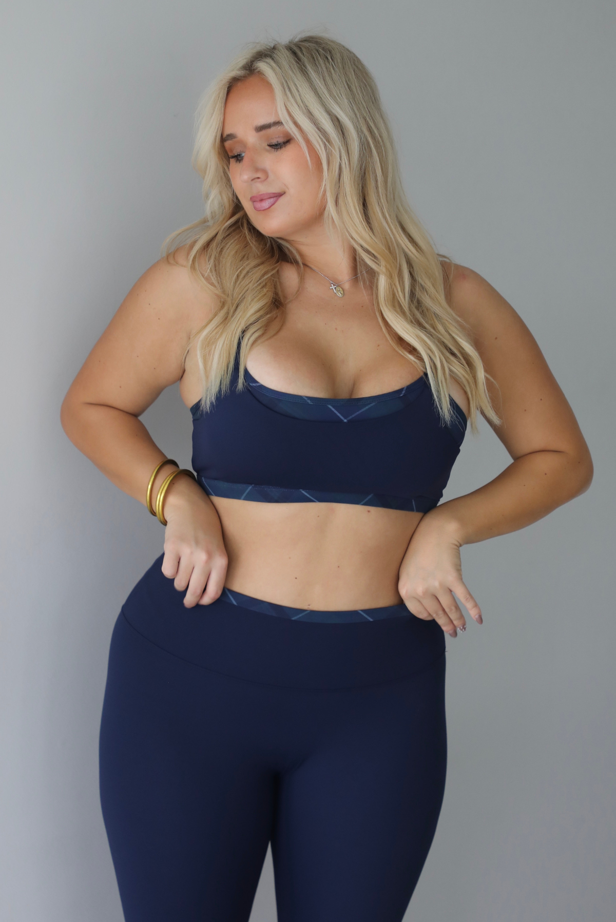 A woman wearing a matching two piece navy athletic set featuring a sports bra and high waisted leggings, standing indoors against a white wall. The photo shows a closeup of the bra, which has plaid trim detailing at the top and bottom, giving it a layered look. 