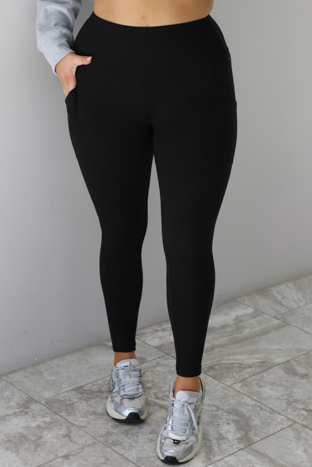 Woman wearing a pair of black leggings. The leggings are high waisted, fitted, with functional pockets on either side. The leggings are worn with a pair of silver sneakers. The photo is taken indoors against a white wall with white marble flooring. 