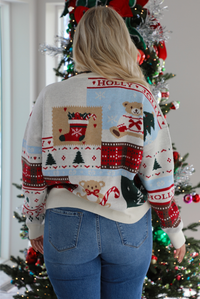Back of a woman model wearing a multi-colored and multi-patterned Christmas sweater with medium wash denim jeans. The photo is taken inside a modern home with a decorated Christmas tree in the back. 