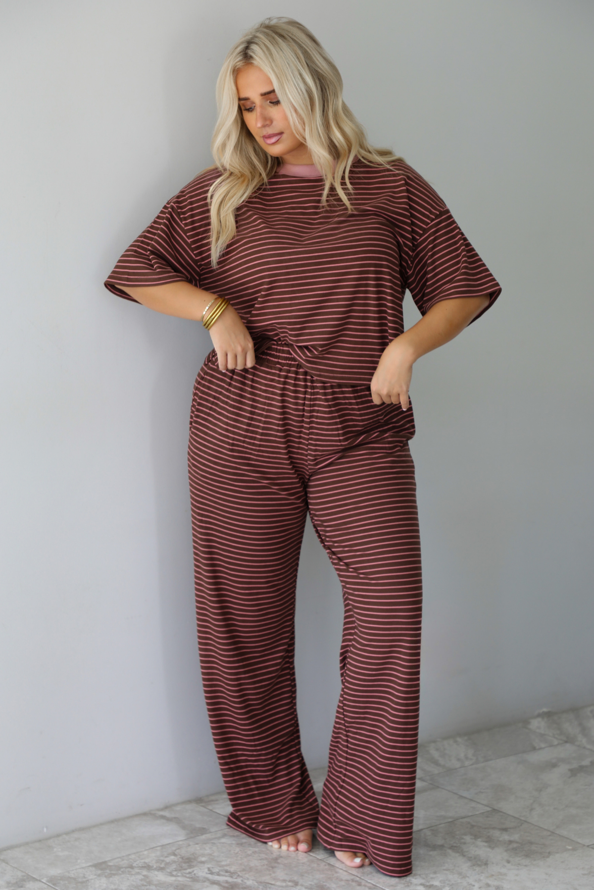 A woman wearing a brown and pink striped two piece set featuring a slightly cropped tee and high waisted pants, with the top slightly tucked into the pants, standing indoors against a white wall with white marble flooring. 
