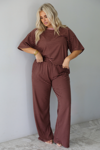 A woman wearing a brown and pink striped two piece set featuring a slightly cropped tee and high waisted pants, with the top slightly tucked into the pants, standing indoors against a white wall with white marble flooring. 