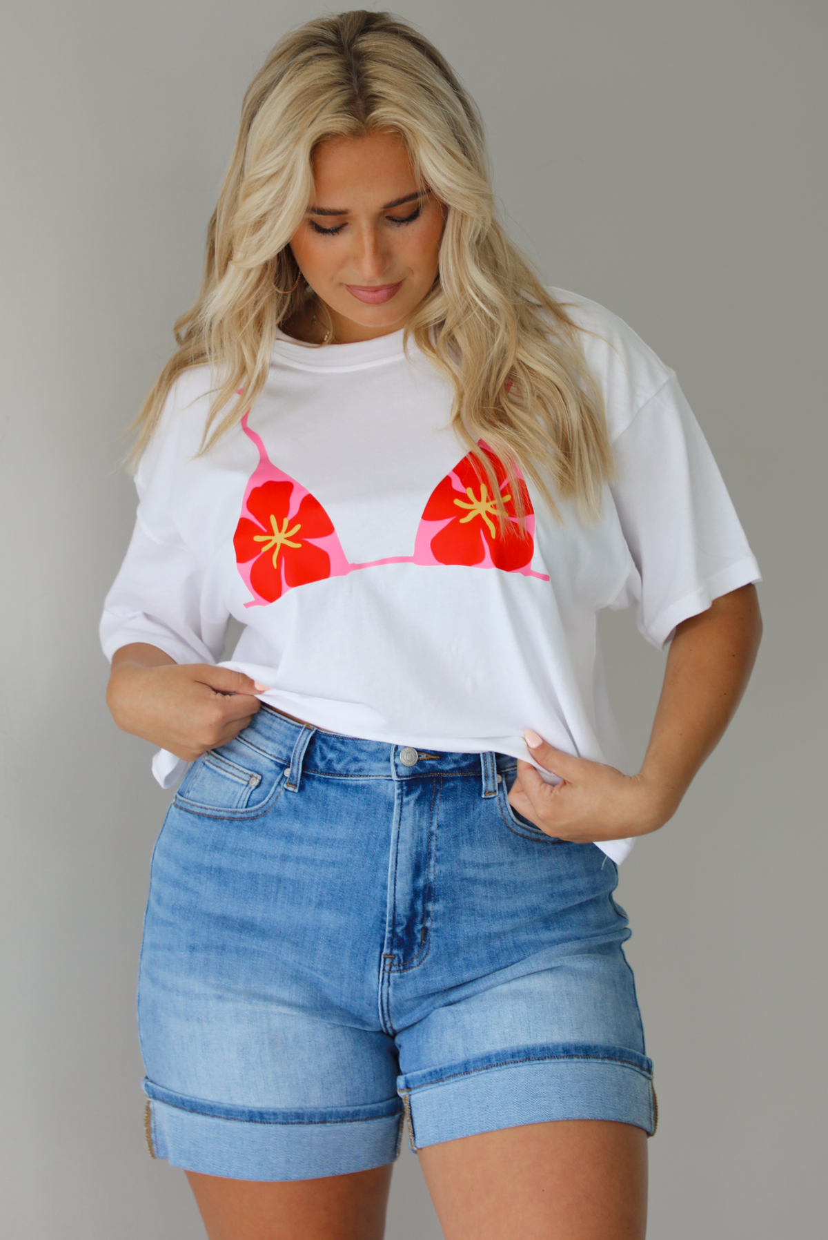 Woman wearing a white cropped boxy tee with a pink, red, and yellow bikini graphic on the front with a pair of medium wash denim shorts indoors against a white wall.