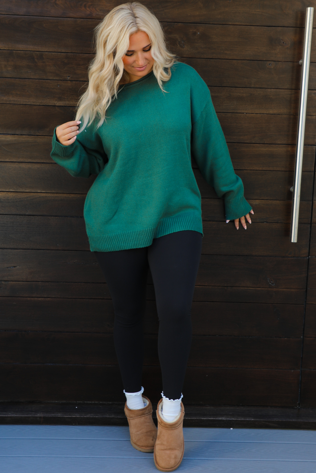 Woman model wearing a green oversized sweater with black leggings, white socks, and tan Uggs. The photo is taken outdoors against a dark brown door. 