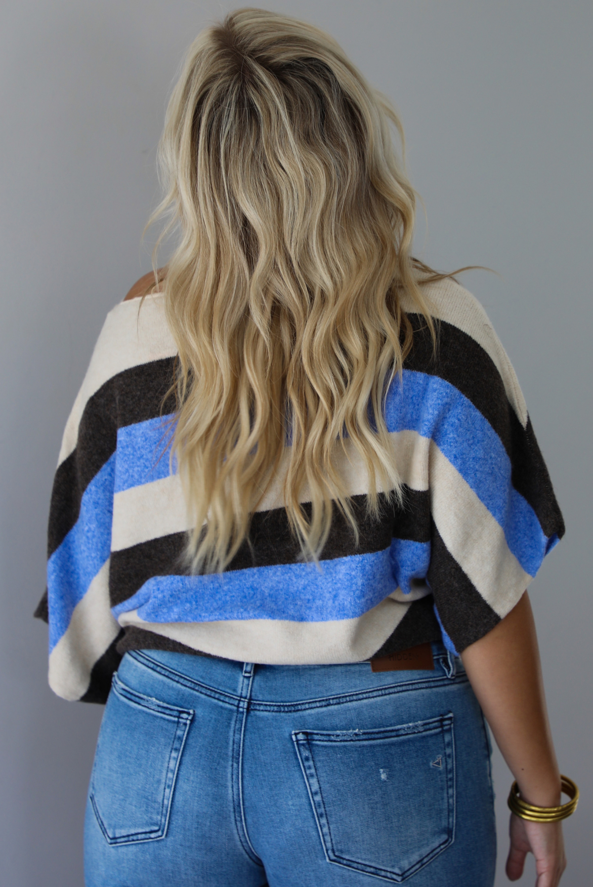 A woman shown from behind wearing a cream, brown,, and blue striped knit top that has a boat-neck design, and quarter length flowy sleeves, paired with a pair of medium wash denim jeans, standing indoors against a white wall.