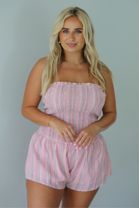 A woman wearing a pink and multi-colored vertically striped strapless romper, standing indoors against a white wall. The photo shows a closeup of the romper, displaying the smocked bodice. 