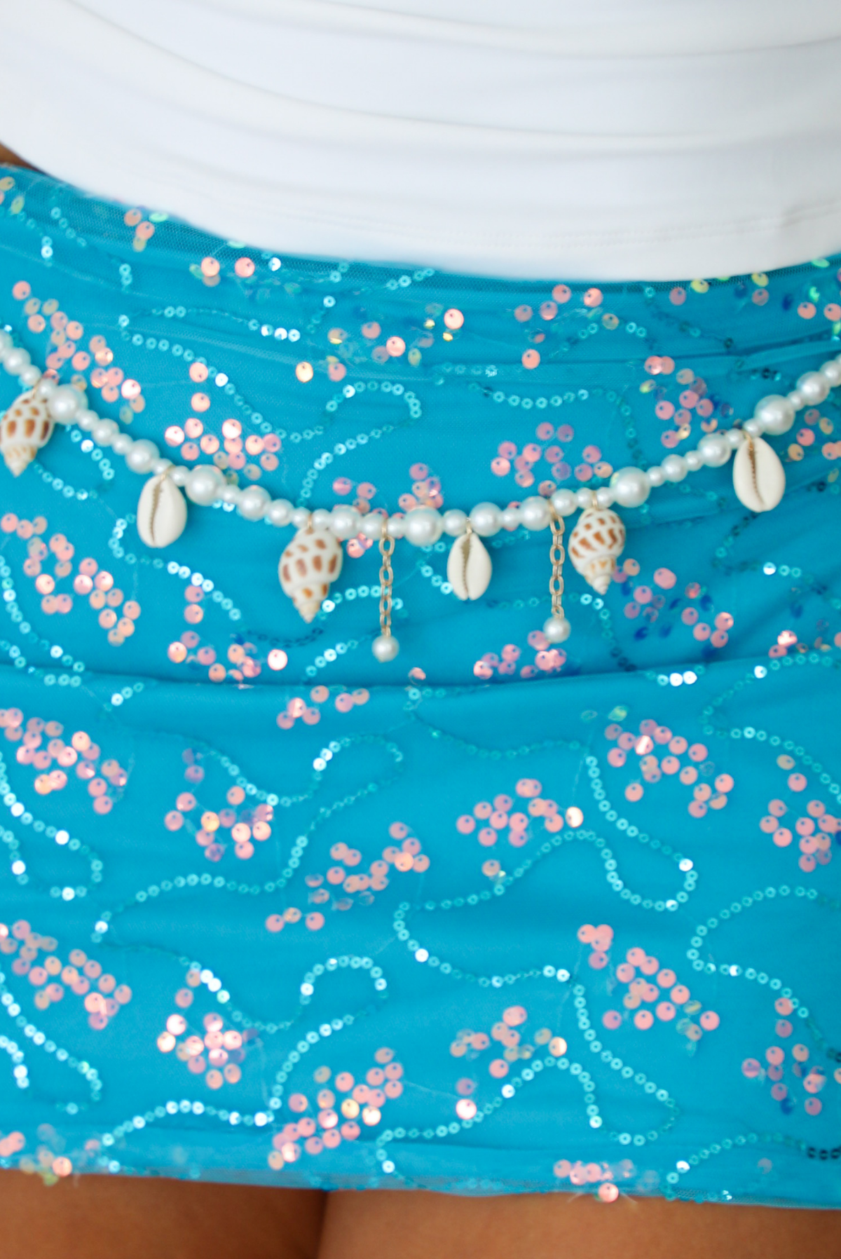 A close-up of a turquoise sequined skirt with pink and silver floral patterns, decorated with a beaded waistband featuring cowrie shells and small dangling shells.