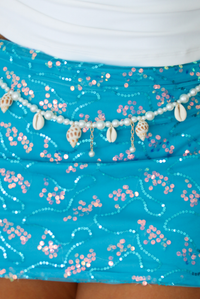 A close-up of a turquoise sequined skirt with pink and silver floral patterns, decorated with a beaded waistband featuring cowrie shells and small dangling shells.