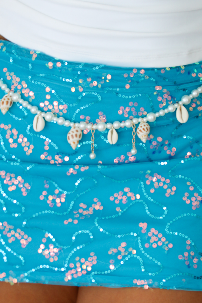 A close-up of a turquoise sequined skirt with pink and silver floral patterns, decorated with a beaded waistband featuring cowrie shells and small dangling shells.