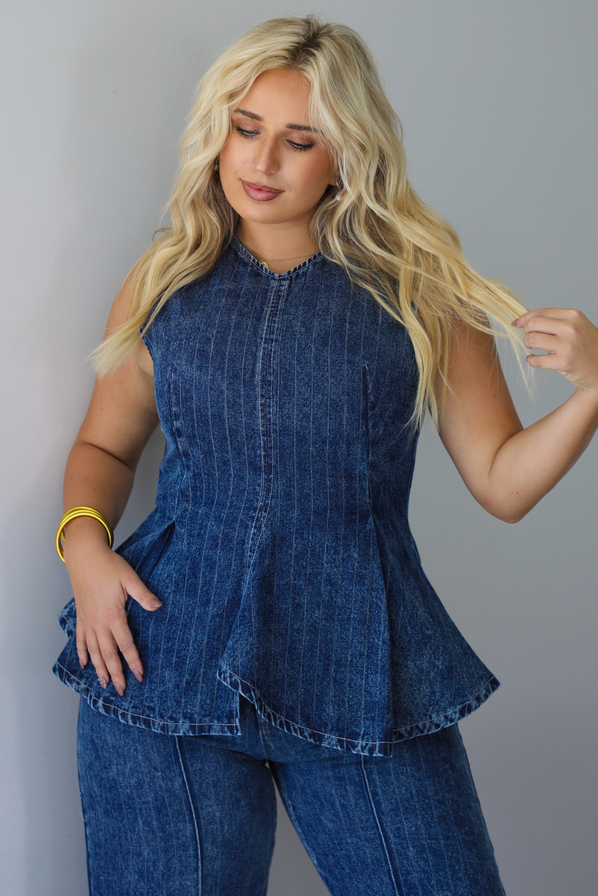 A woman wearing a matching two piece dark wash denim set with a pin striped design on it featuring a sleeveless peplum style top and high waisted denim jeans, standing indoors against a white wall.