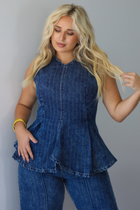 A woman wearing a matching two piece dark wash denim set with a pin striped design on it featuring a sleeveless peplum style top and high waisted denim jeans, standing indoors against a white wall.