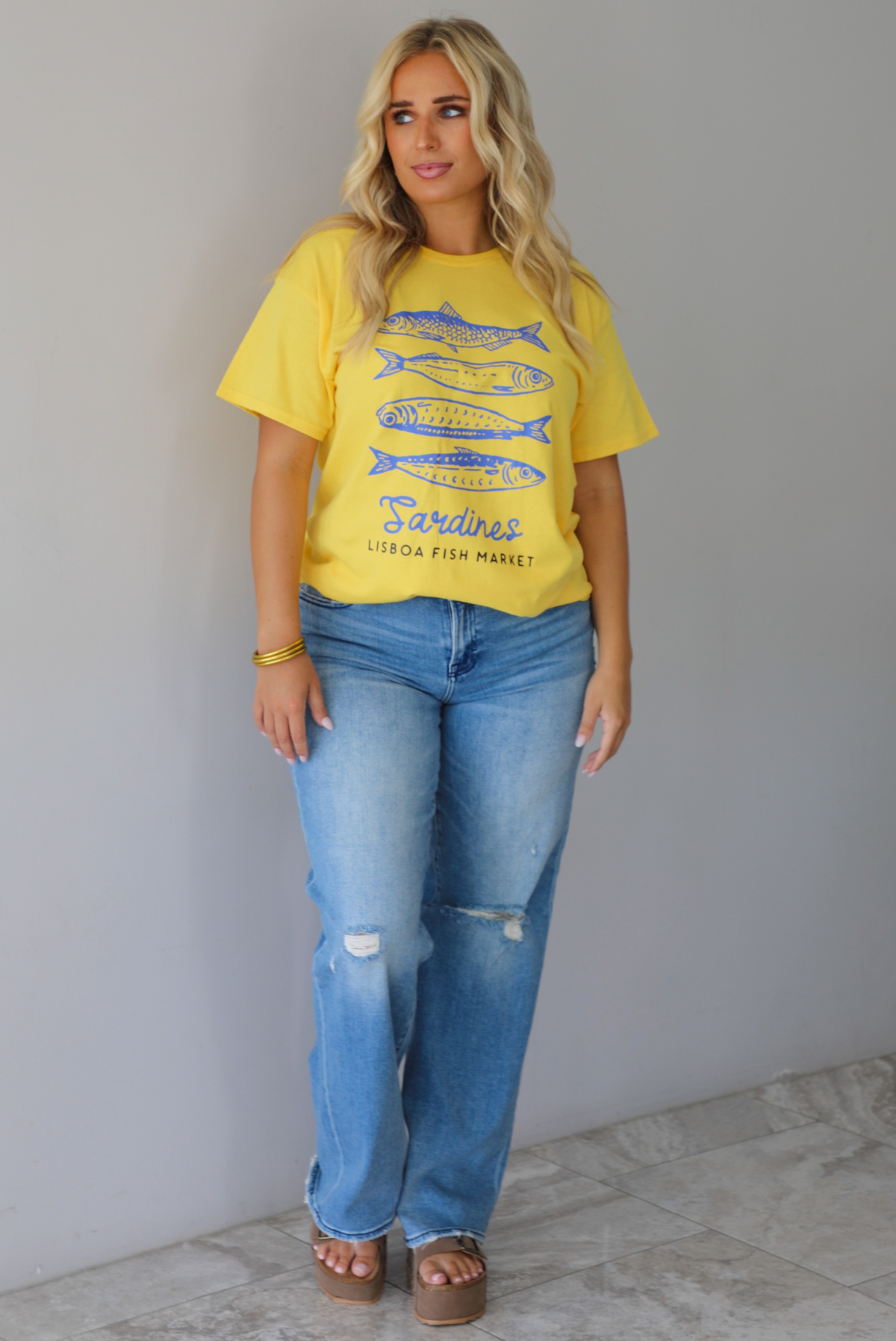 A woman wearing a bright yellow graphic t-shirt with blue sardine illustrations and the text 'Sardines LISBON FISH MARKET', paired with blue jeans, standing indoors against a white wall with white marble flooring. 