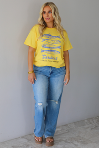 A woman wearing a bright yellow graphic t-shirt with blue sardine illustrations and the text 'Sardines LISBON FISH MARKET', paired with blue jeans, standing indoors against a white wall with white marble flooring. 