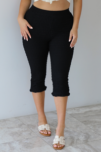 Closeup of a woman wearing high waisted black capri pants that have a textured feel to them and a slight ruffled hem indoors against a white wall with white marble flooring. 