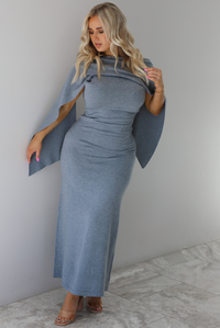 Woman wearing a grey maxi length knit dress that has a high neckline with an attached shawl detail that covers the upper half of the chest and flows on either shoulder in the back. The dress is fitted with a slightly flowy lower hem. The dress is paired with clear strapped nude heels. The photo is taken indoors against a white wall with white marble flooring. 