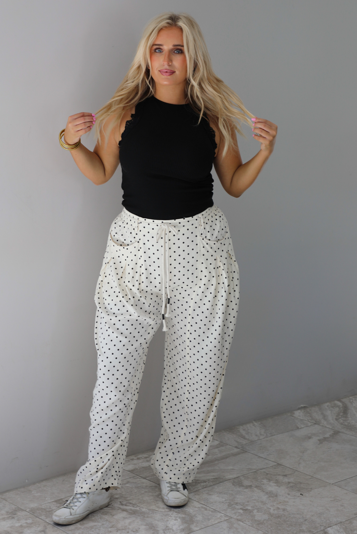 A woman wearing a black high neck tank paired with sand white pants with a black polka dot print pattern throughout, standing indoors against a white wall with white marble flooring. 