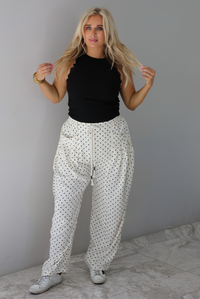 A woman wearing a black high neck tank paired with sand white pants with a black polka dot print pattern throughout, standing indoors against a white wall with white marble flooring. 