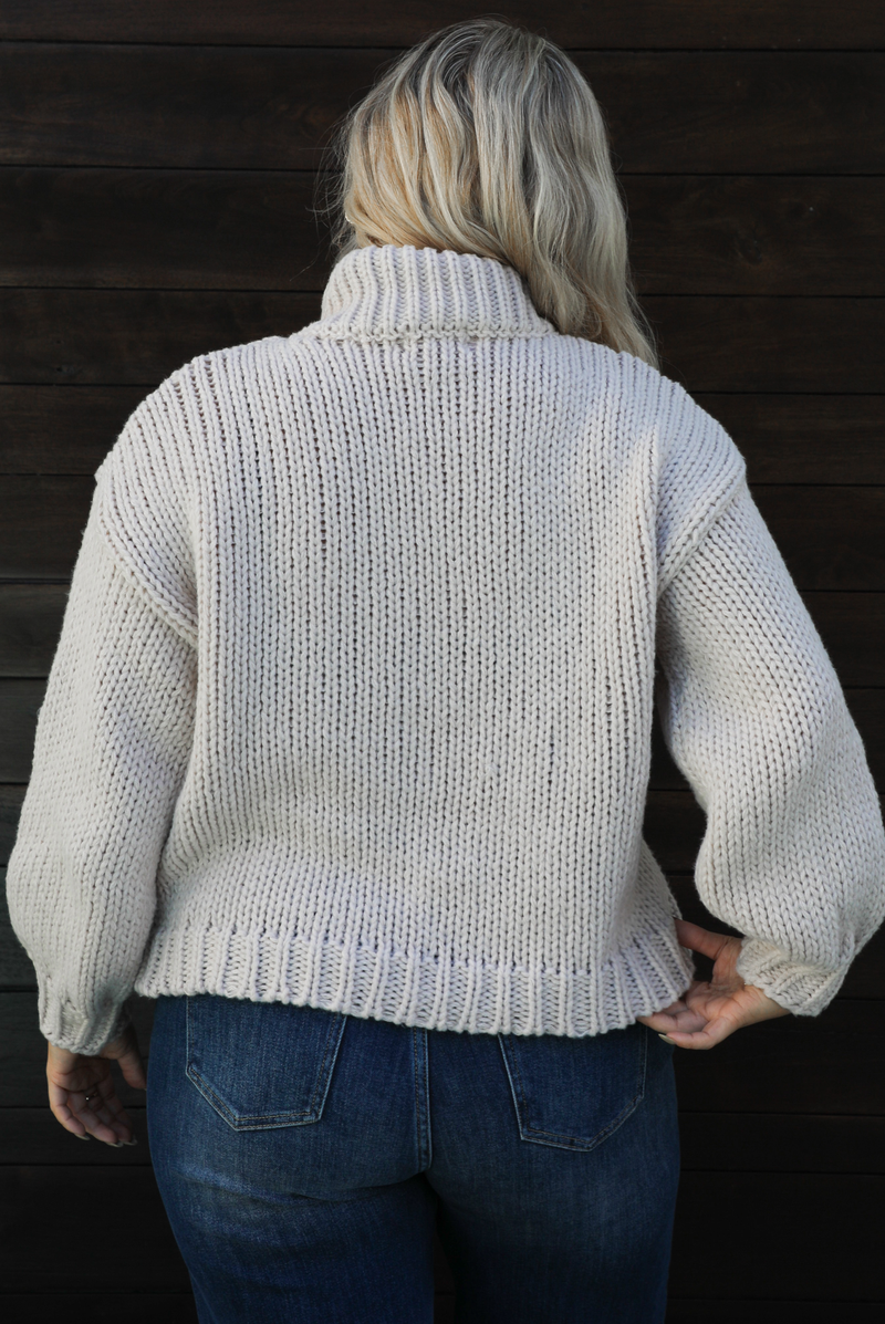 Close up of the back of a woman model wearing a chunky knit turtle neck sweater with dark denim jeans outdoors in front of a dark wood door. 