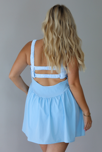 A woman shown from the back wearing a light blue sleeveless dress with a ruffled open back and wide shoulder straps that are a blue and white gingham print, standing indoors against a white wall. 
