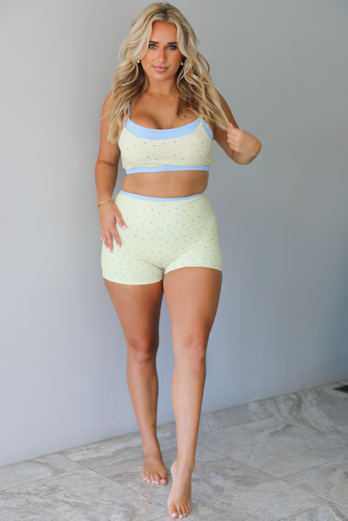 Woman wearing a yellow and blue two piece athletic set featuring a sports bra and high waisted biker shorts with a light blue small floral print pattern throughout, while standing indoors against a white wall with white marble flooring. 