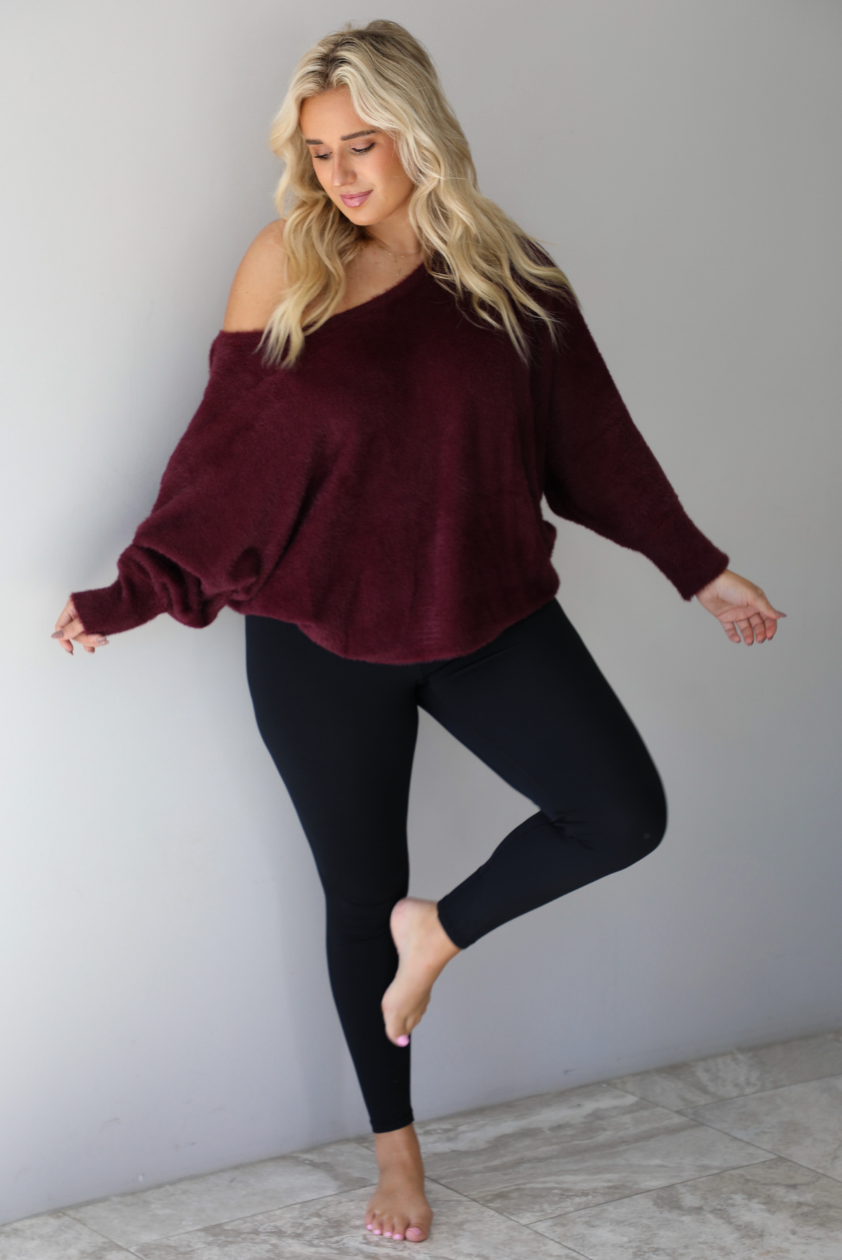 A woman wearing a maroon oversized long sleeved sweater, worn off the shoulder, paired with black leggings, indoors against a white wall with white marble flooring.