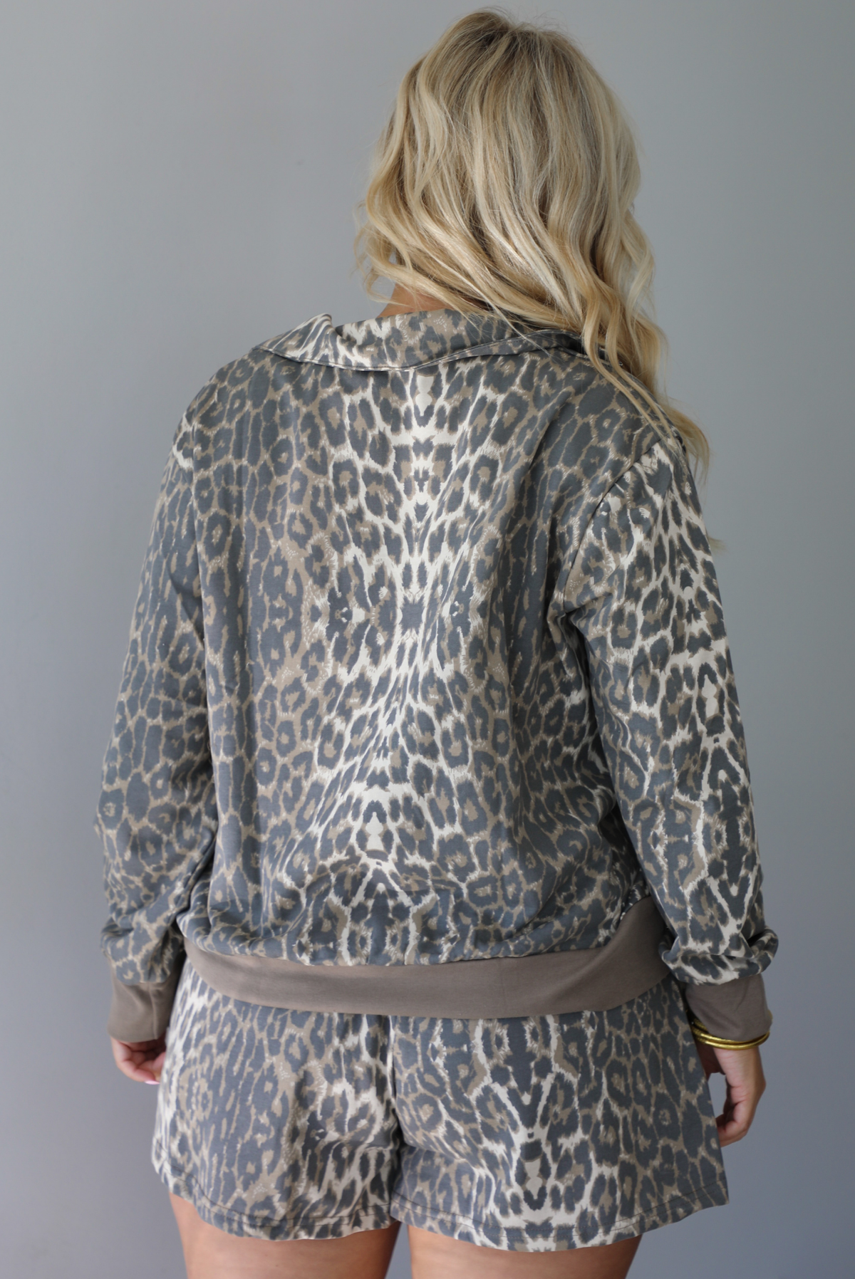 A woman shown from behind modeling a matching leopard-print quarter-zip sweater, untucked to show the full length, and shorts set with gray drawstrings, standing indoors against a white wall.