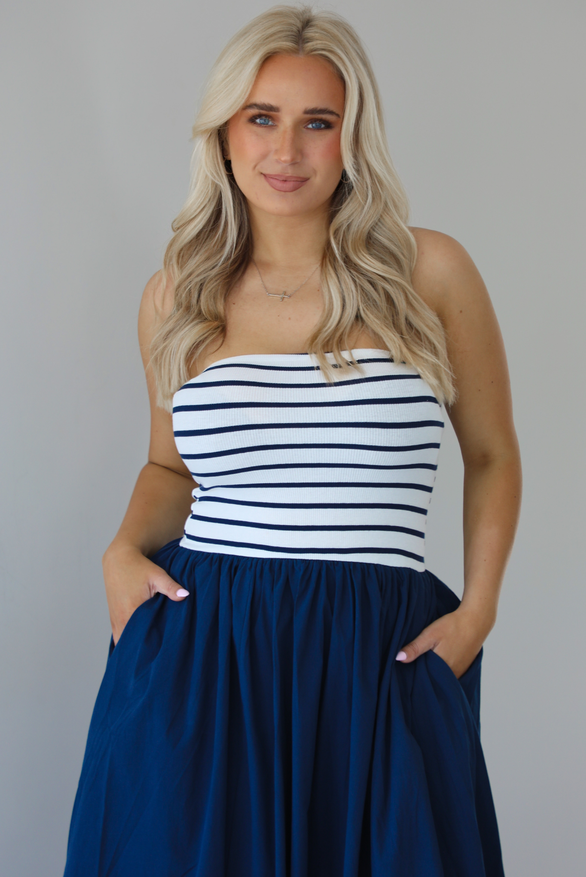 Woman wearing a maxi dress featuring a navy and white striped bodice and navy, maxi length skirt. The photo is taken indoors against a white wall and shows a closeup of the dress with functional pockets on either side. 
