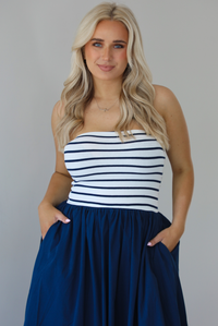 Woman wearing a maxi dress featuring a navy and white striped bodice and navy, maxi length skirt. The photo is taken indoors against a white wall and shows a closeup of the dress with functional pockets on either side. 