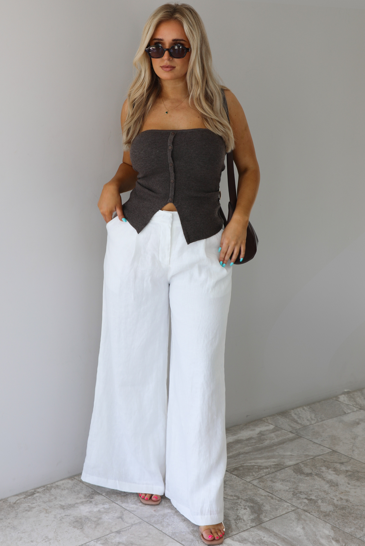 Woman wearing a strapless brown top with non-functional button closures in the front with  a split hem detail, and high waisted, flared hem white linen pants with a brown shoulder bag. The photo is taken indoors against a white wall with white marble flooring. 
