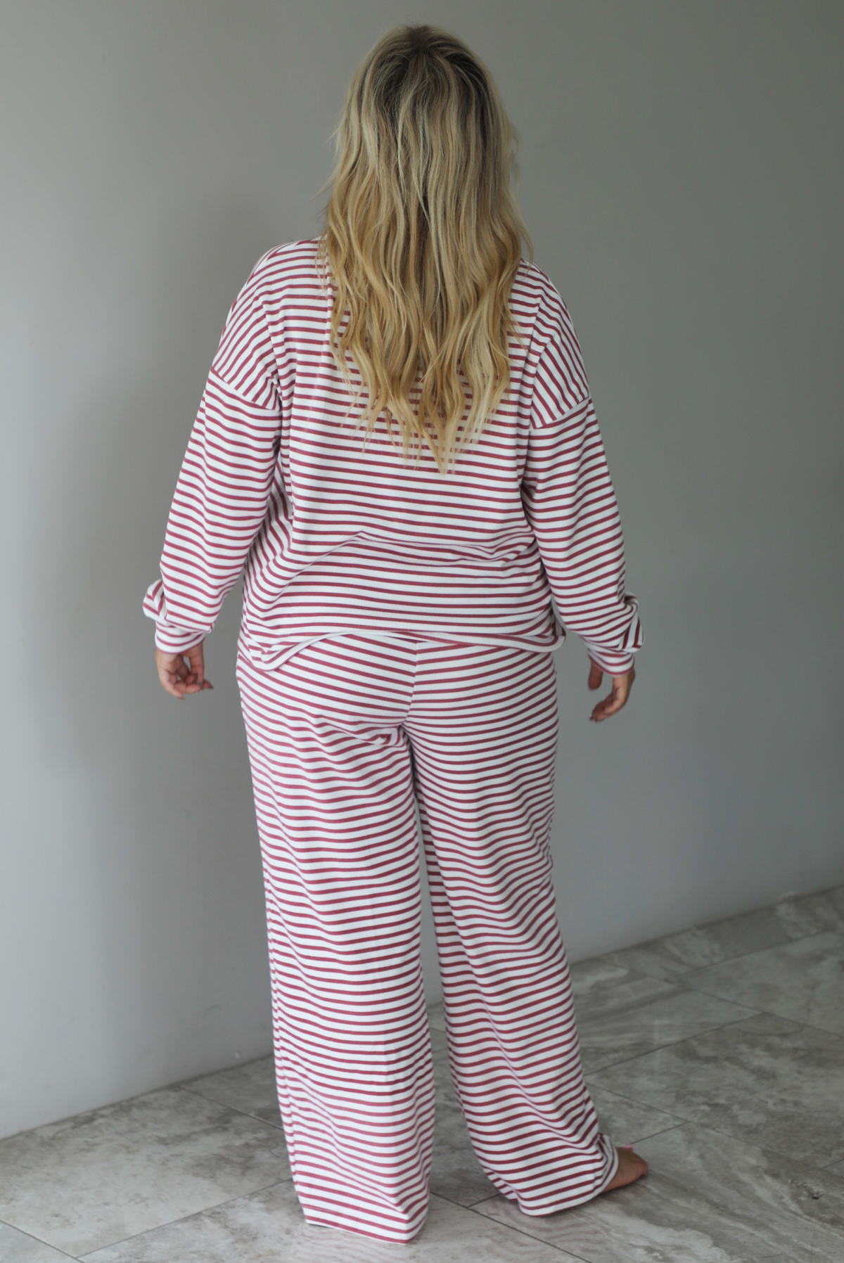 A woman shown from behind wearing a matching two piece set featuring a long sleeved top and high waisted wide leg pants with a berry and white striped pattern throughout, standing indoors against a white wall with white marble flooring. 