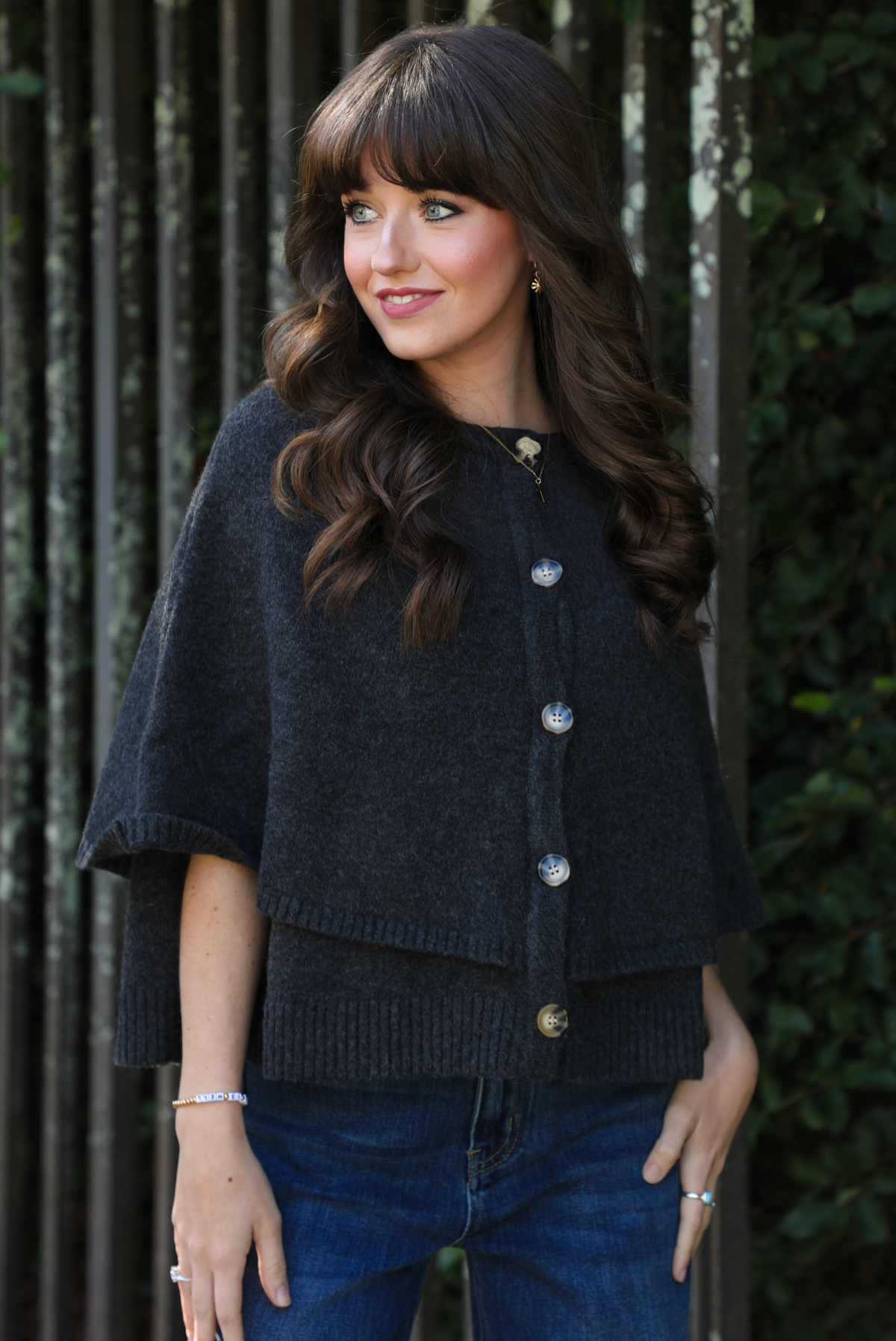 Close up of a woman model outdoors wearing a charcoal grey colored cape cardigan with functional button closures in the front and dark wash denim. 