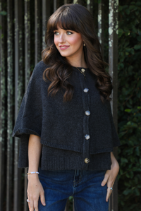 Close up of a woman model outdoors wearing a charcoal grey colored cape cardigan with functional button closures in the front and dark wash denim. 