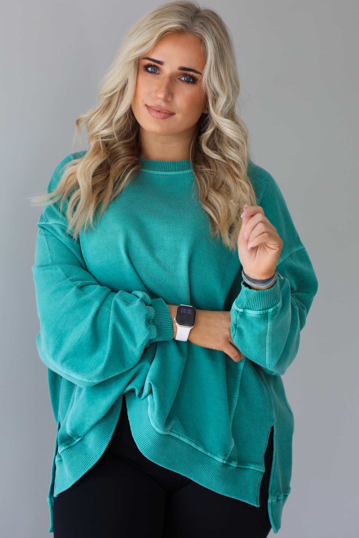 Woman wearing a teal oversized sweater with black leggings that have a slightly flared hem with split hem details in the front. The photo shows a closeup of the sweater and has a white background. 