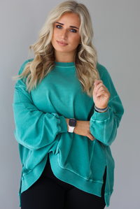 Woman wearing a teal oversized sweater with black leggings that have a slightly flared hem with split hem details in the front. The photo shows a closeup of the sweater and has a white background. 