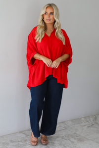 A woman wearing a bright red oversized short-sleeve blouse and dark wide-leg jeans, standing indoors against a white wall with white marble flooring. 