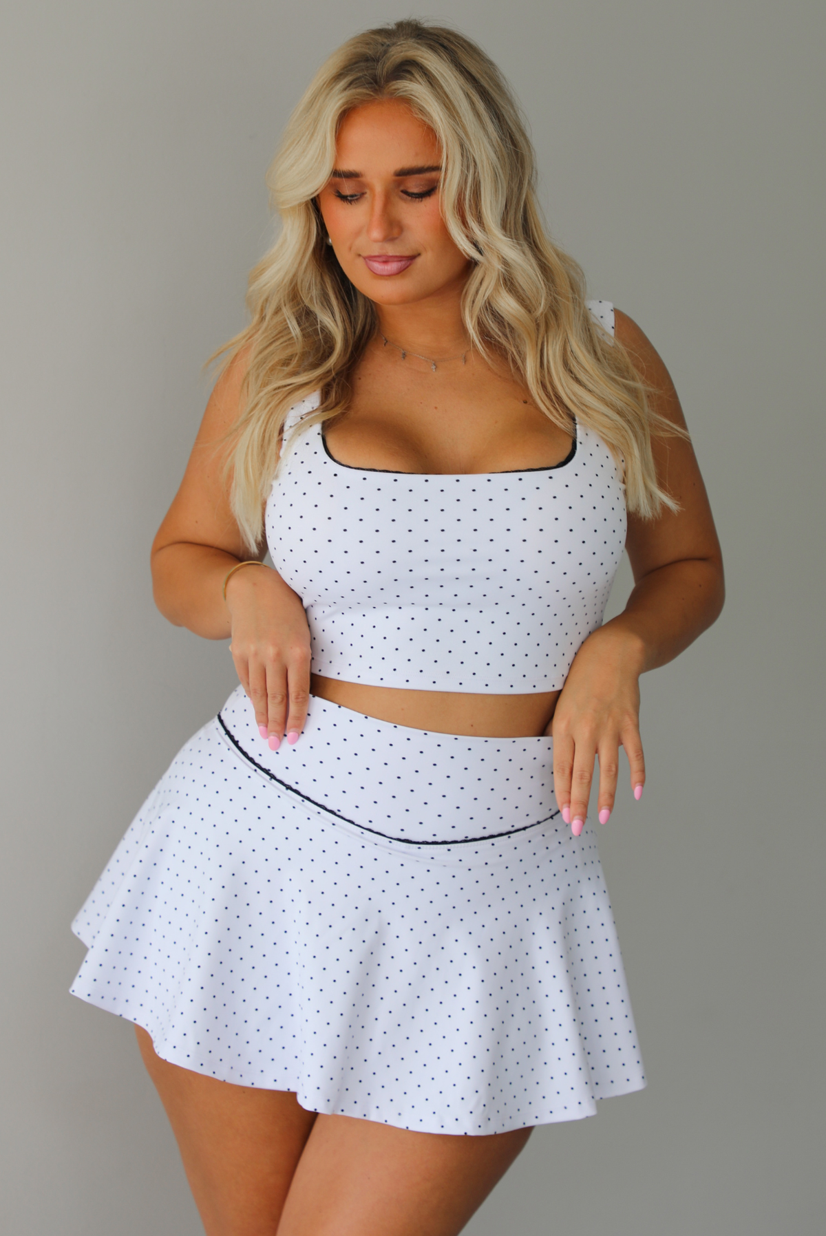A woman modeling a white two-piece lounge set with small black polka dots, featuring a scoop-neck crop top and a matching flared skirt, standing indoors against a white wall.