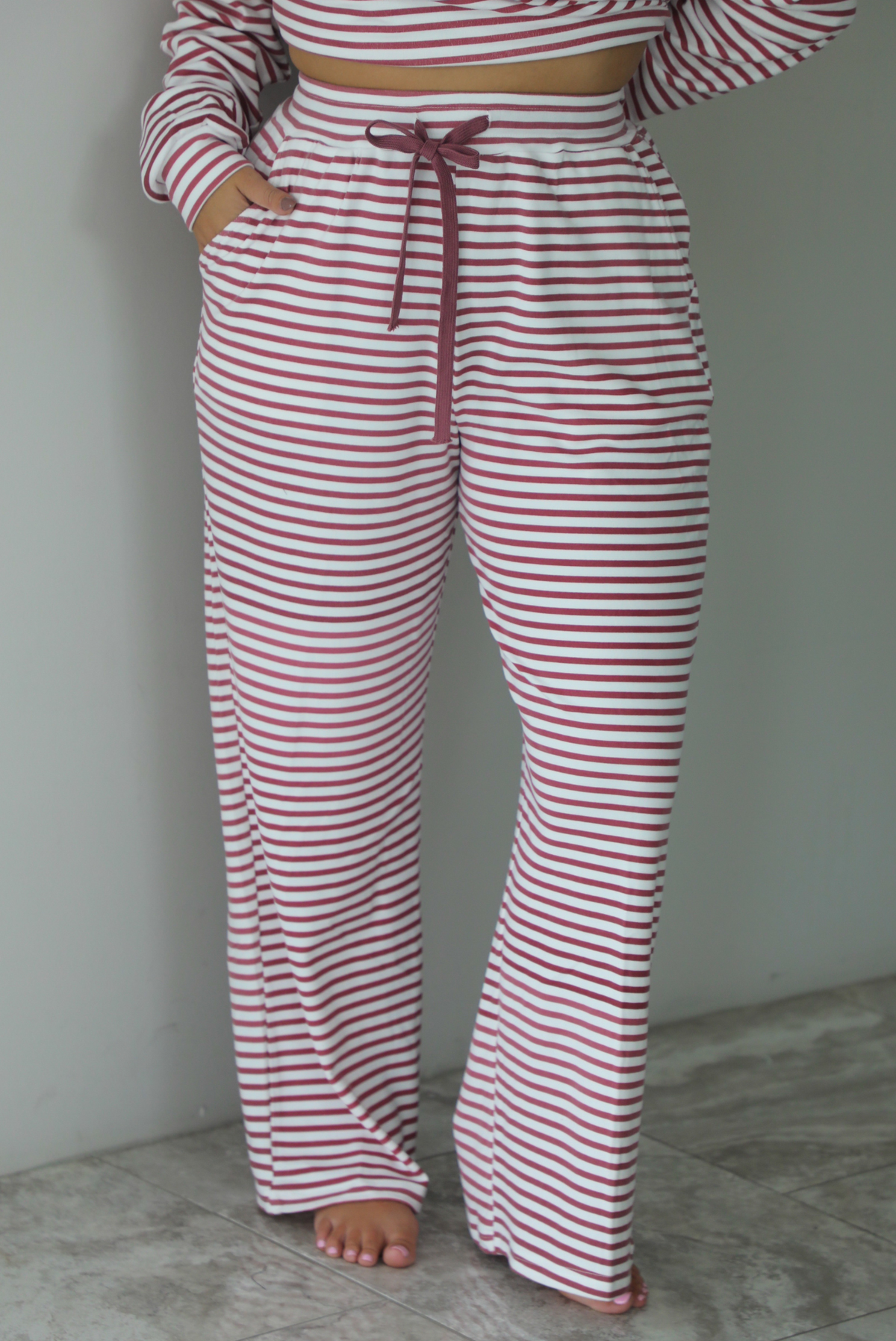 A woman wearing a matching two piece set featuring a long sleeved top and high waisted wide leg pants with a berry and white striped pattern throughout, standing indoors against a white wall with white marble flooring. The photo shows a closeup of the pants, which have an elastic and drawstring waistline and functional pockets on either side. 