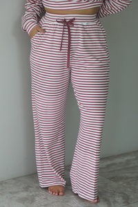 A woman wearing a matching two piece set featuring a long sleeved top and high waisted wide leg pants with a berry and white striped pattern throughout, standing indoors against a white wall with white marble flooring. The photo shows a closeup of the pants, which have an elastic and drawstring waistline and functional pockets on either side. 