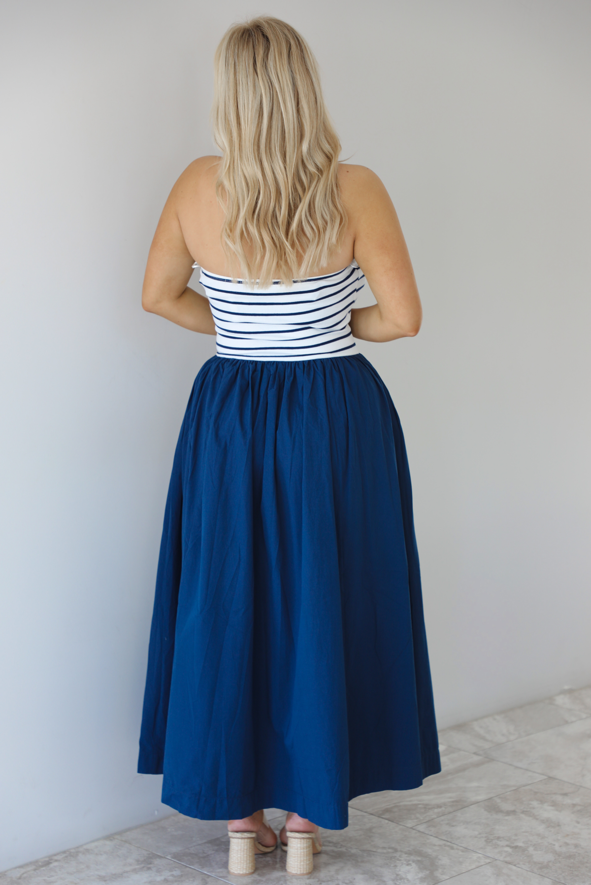 Woman wearing a maxi dress featuring a navy and white striped bodice and navy, maxi length skirt. The photo is taken indoors against a white wall with white marble flooring and shows the back of the dress. 