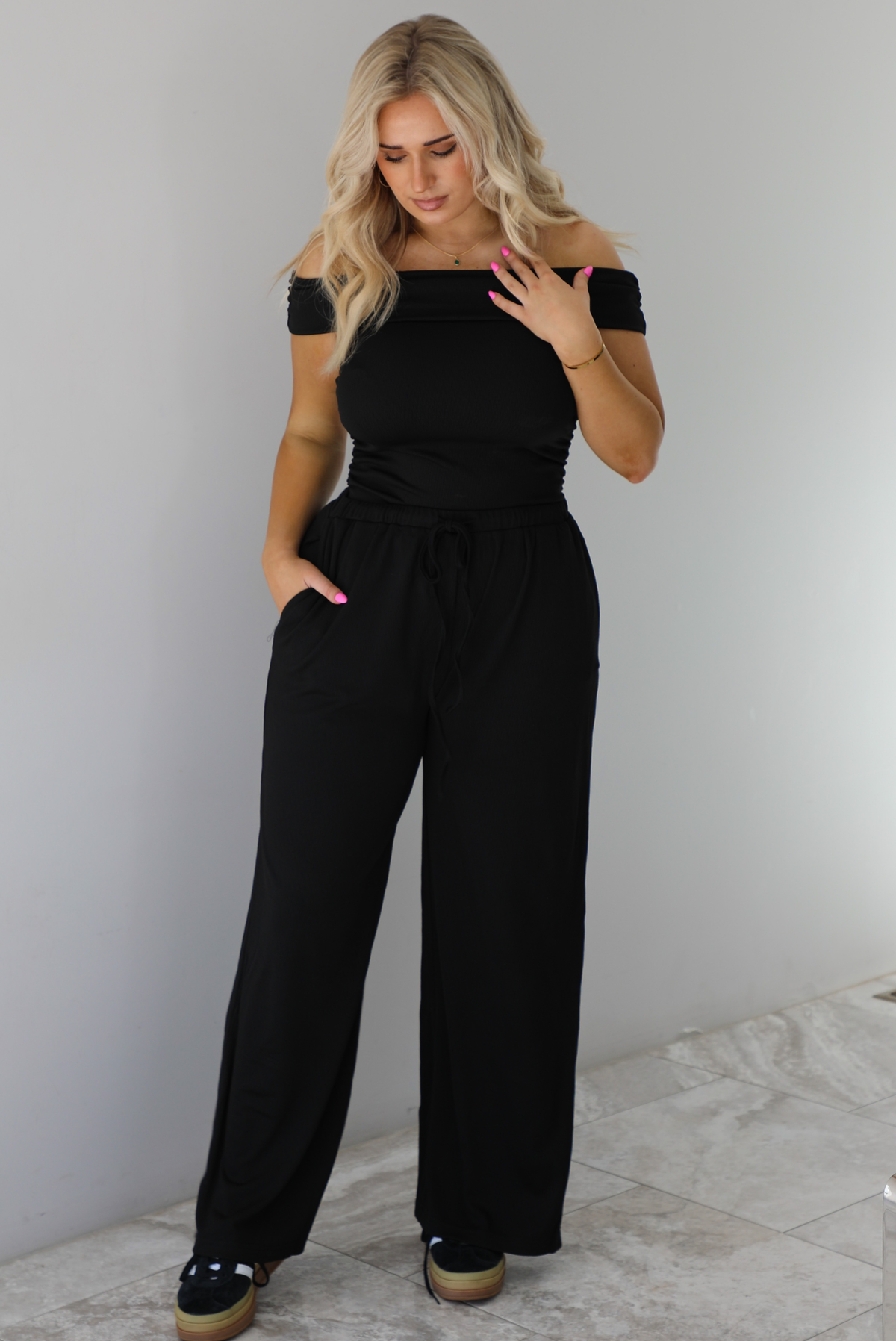 Woman wearing a two piece set featuring an off the shoulder top and high waisted pants. The photo is taken indoors against a white wall with white marble flooring. 