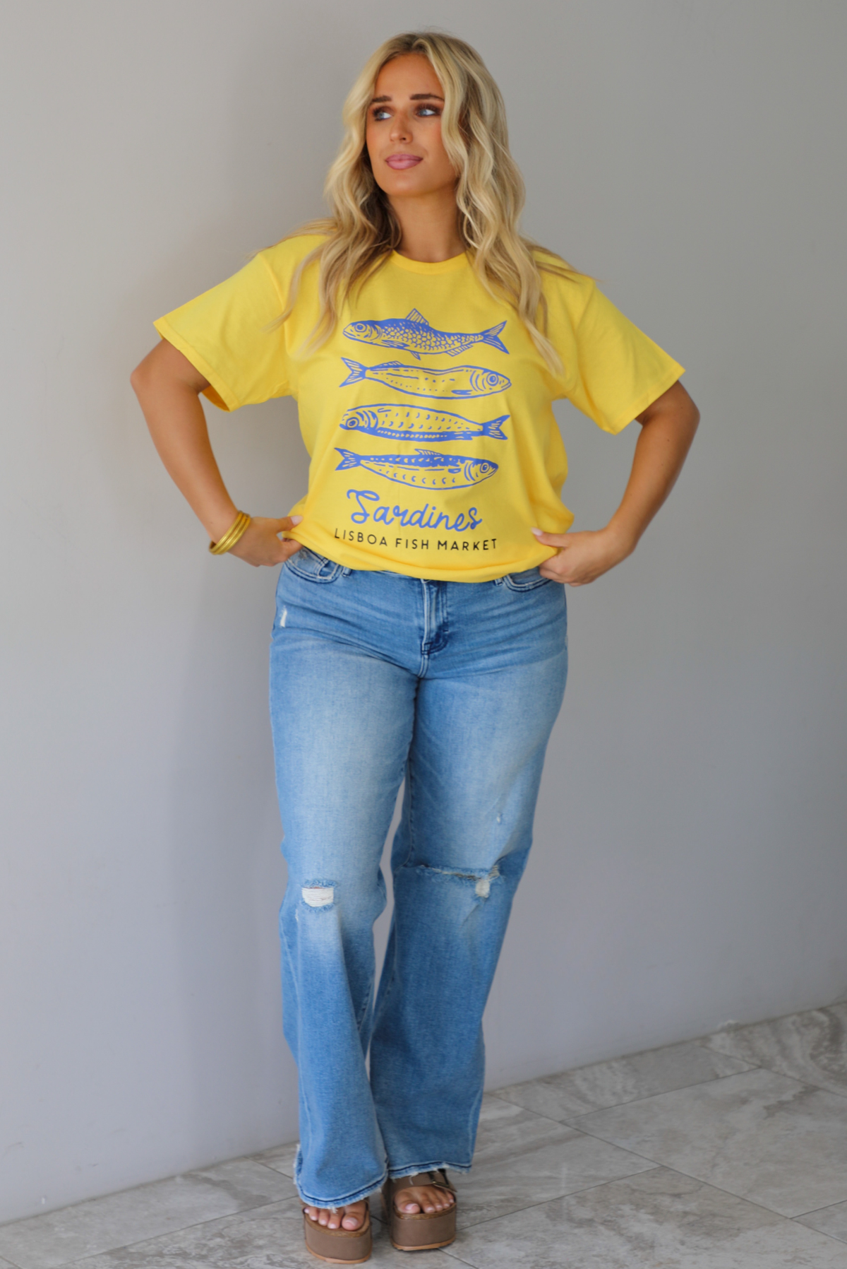 A woman wearing a bright yellow graphic t-shirt with blue sardine illustrations and the text 'Sardines LISBON FISH MARKET', paired with blue jeans, standing indoors against a white wall with white marble flooring. 