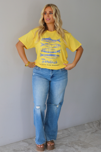 A woman wearing a bright yellow graphic t-shirt with blue sardine illustrations and the text 'Sardines LISBON FISH MARKET', paired with blue jeans, standing indoors against a white wall with white marble flooring. 