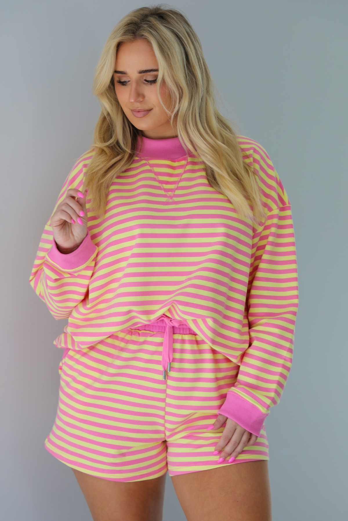 Woman wearing a pink and yellow striped set featuring an oversized sweatshirt and shorts. The photo is taken indoors against a white wall and shows a closeup of the set, which shows the top tucked into the front of the shorts. 