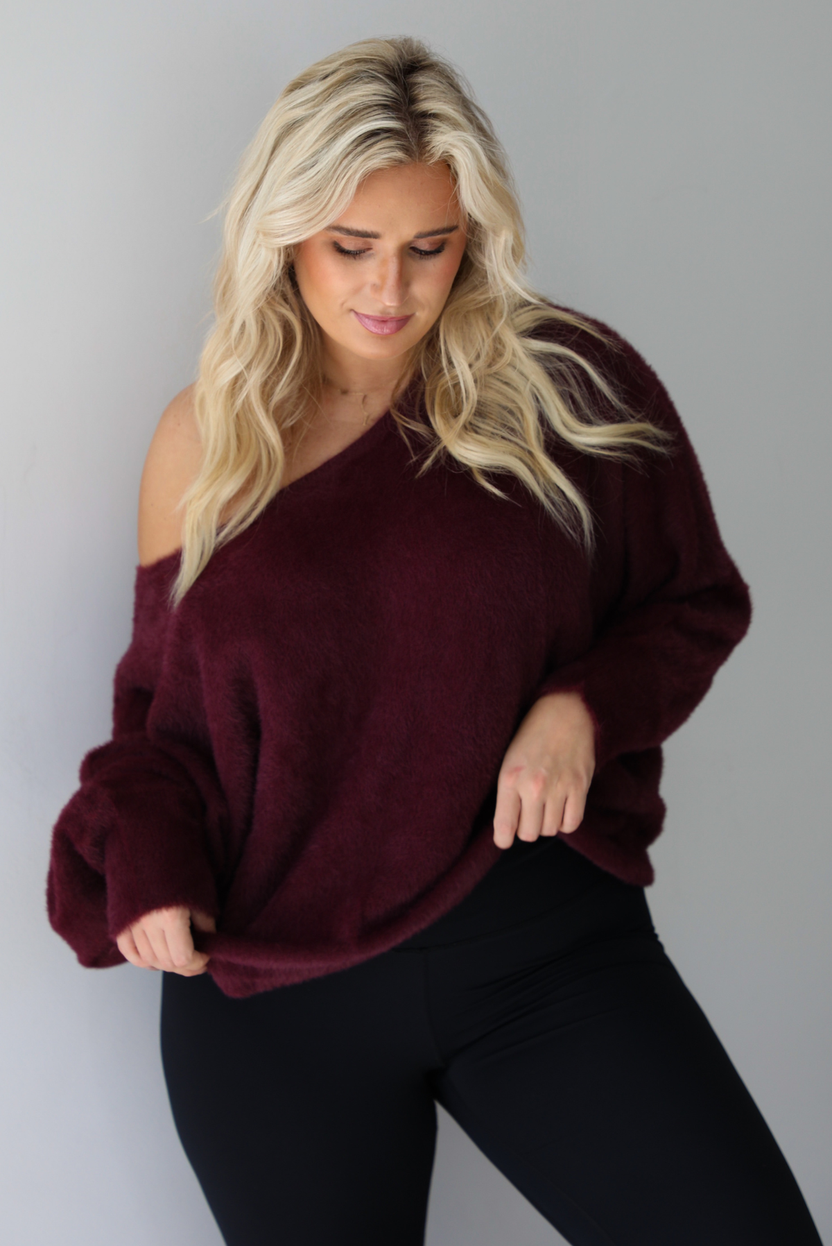A woman wearing a maroon oversized long sleeved sweater, worn off the shoulder, paired with black leggings, indoors against a white wall.