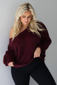 A woman wearing a maroon oversized long sleeved sweater, worn off the shoulder, paired with black leggings, indoors against a white wall.